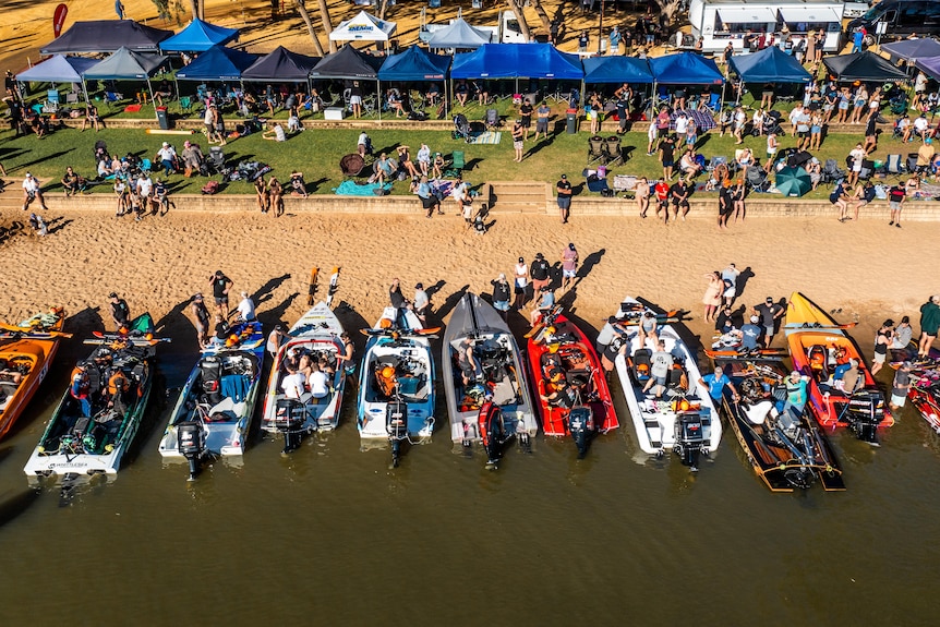 13 water ski boats pointing at a sandbar, with people sitting on nearby grass and under blue gazebos