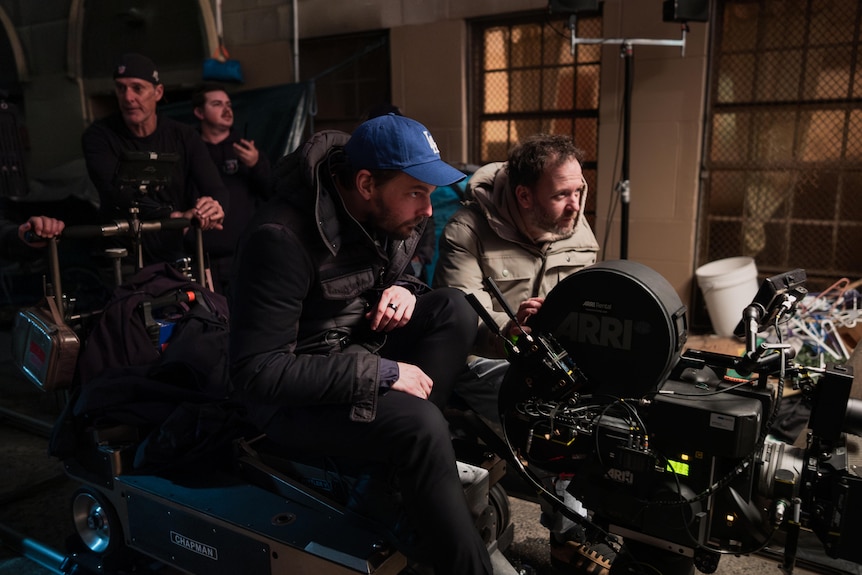 Two men sit in a camera rig while two more men sit behind them.