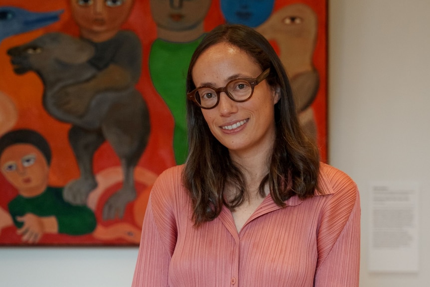 A woman with long brown hair and glasses, wearing a pink buttoned-up cardigan, standing in front of a painting in a gallery.