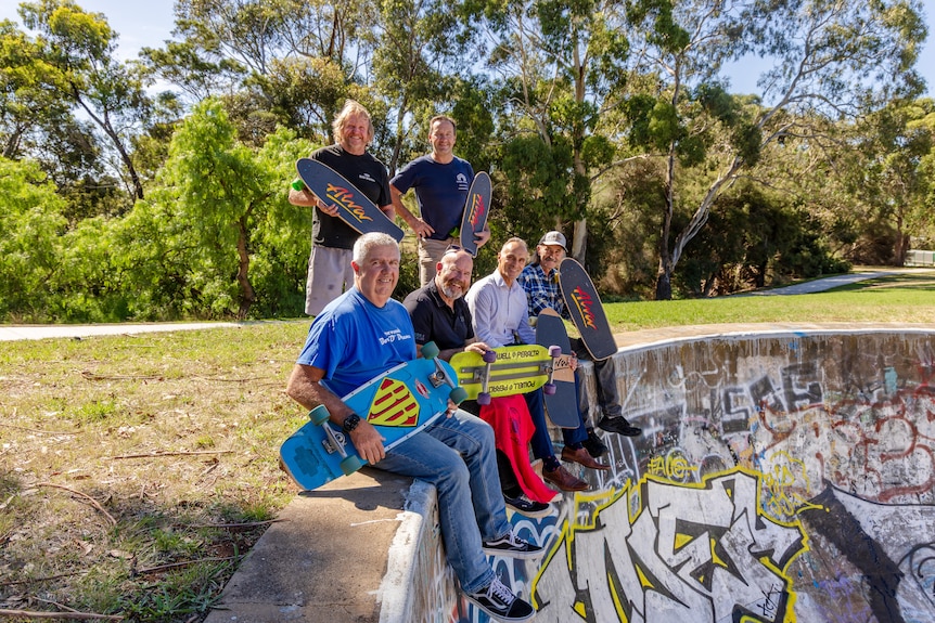 A group of older skaters sits together at the side of the concrete bowl.