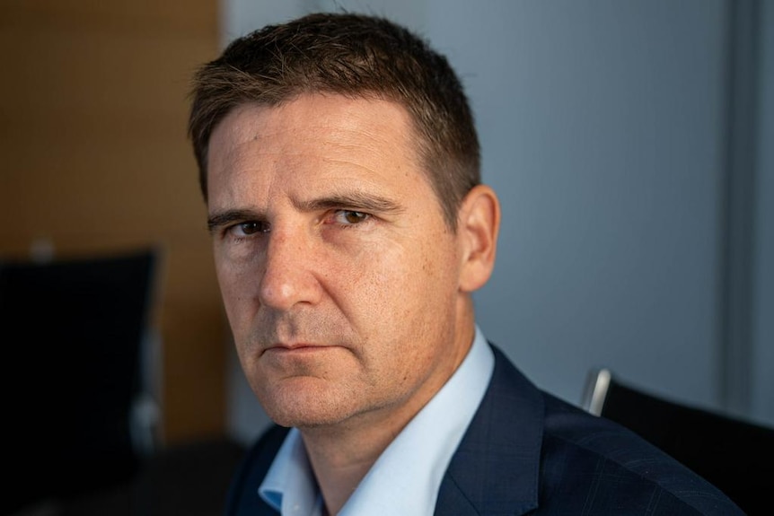 A dark-haired man in a dark blazer stands in an office.