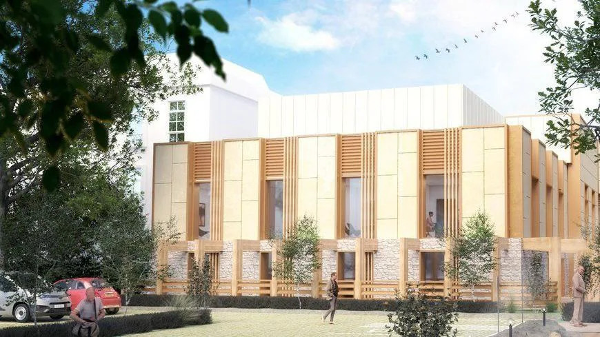 An artist's impression of a modern cancer treatment centre with cream cladding, light wooden details and large windows. There is a large green space and a car park to the front, with trees surrounding.