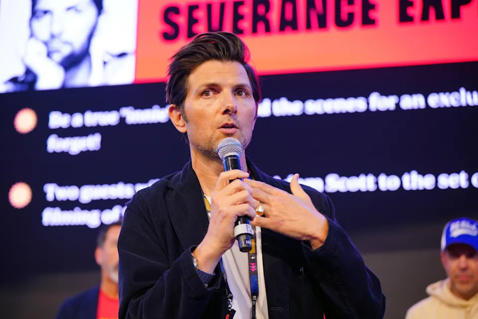 Adam Scott speaking at an event, holding a microphone, dressed in a casual blazer