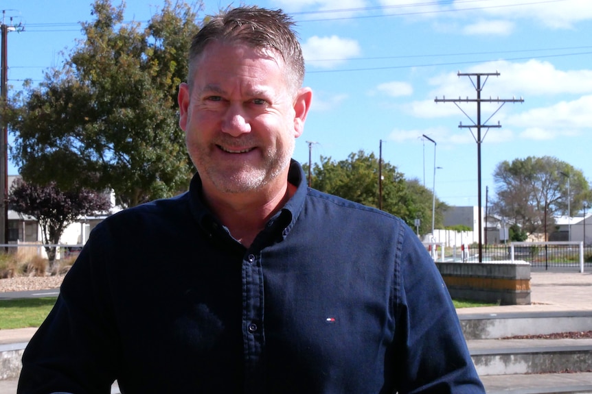 A man with brown-grey hair wearing a dark blue collared shirt stands near an abandoned rail turntable on a sunny day.