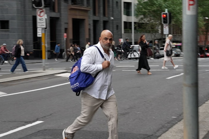 A man in a white shirt, beige pants and carrying a blue backpack crosses a city street.