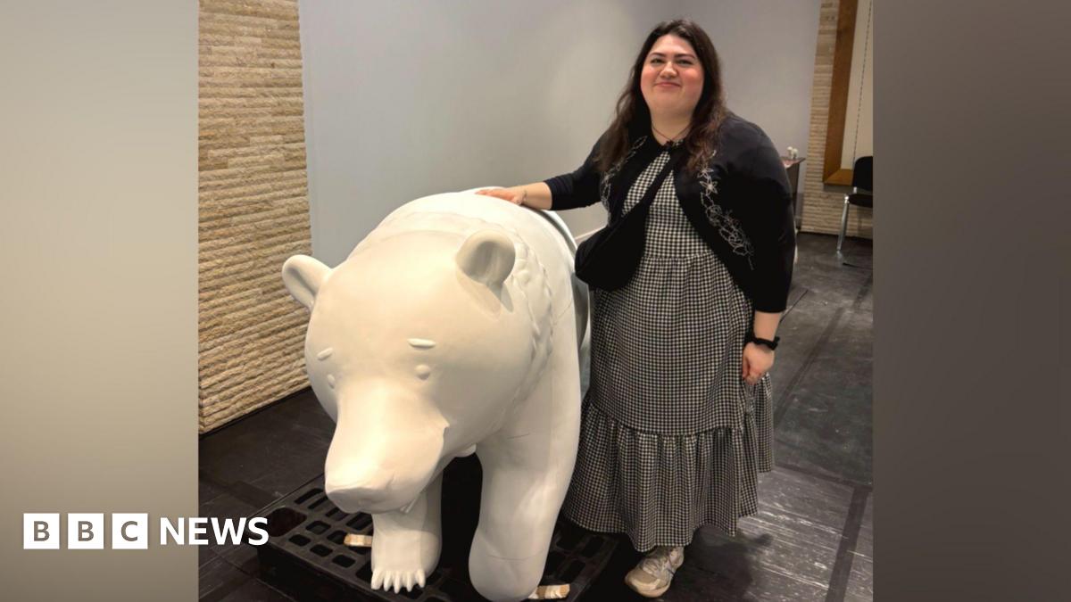 Becky Shepperson in a long dress and cardigan, smiling with her hand on a bear sculpture. The bear is white as it has not been painted yet.