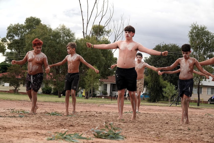 A young man dances on red dirt with a group of others. On his face and body is white paint