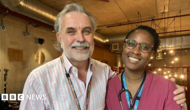 Ashley Brown is seen in a pink and white striped shirt with a rainbow NHS lanyard stands next to Mainga Bhima in burgundy medical scrubs with a stethoscope around her neck. They are both smiling in an indoor setting with industrial-style lighting in the background.