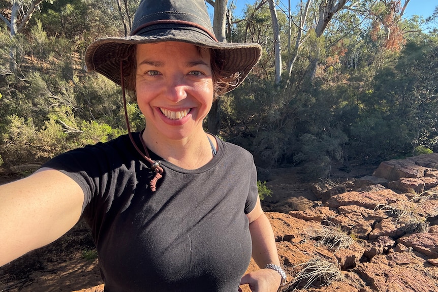 Selfie of a woman in the bush on a rock wearing a black t-shirt and broad-brimmed hat smiling
