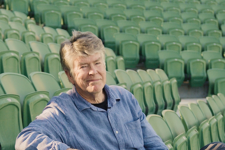 A middle-aged man sits in a grandstand, holding a black sports helmet.