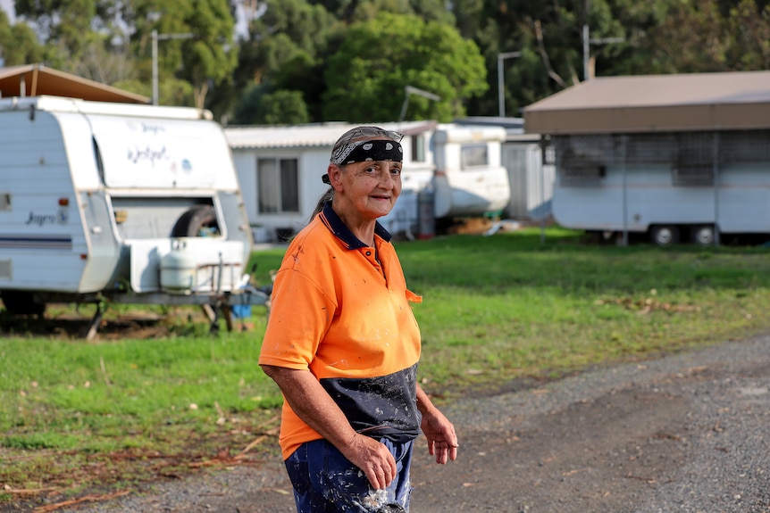 Older woman wearing orange high vis shirt, blue work pants and black bandana stands in country caravan park