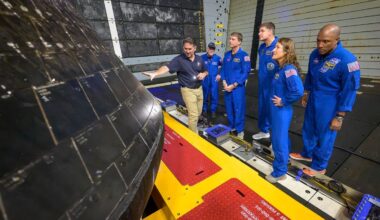 six people, including four nasa astronauts wearing blue flight suits, inspect the black bottom region of a conical space capsule inside a ship's well deck at sea