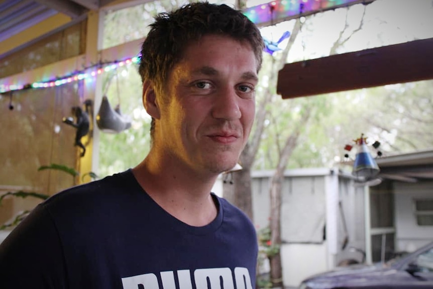 A man in a blue shirt looks at the camera. Behind him, lights hang from a rafter.