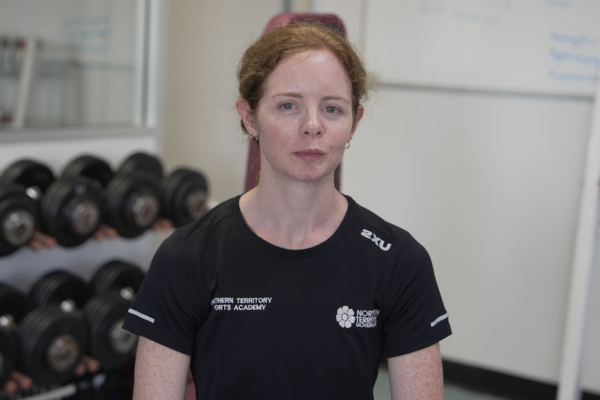 A woman looking at the camera with a serious facial expression as she sits in front of gym equipment.