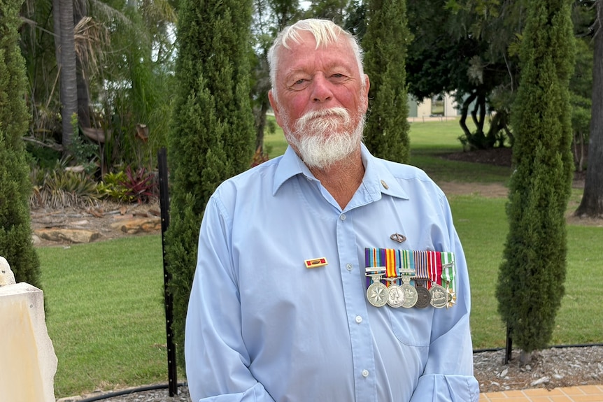 A man with white hair and beard with a row of medals on his chest standing in front of pine trees.