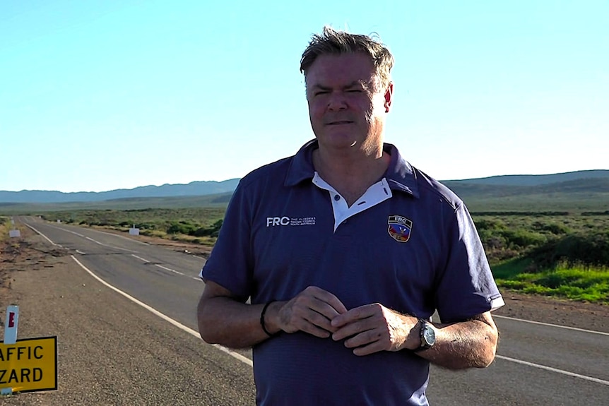 A man stands in front of the flinders ranges, a road sign is seen in the background.