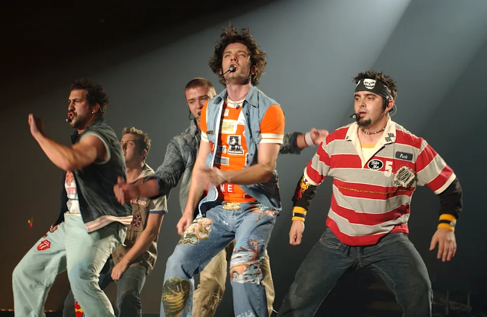 Five performers on stage, mid-dance, wearing casual, eclectic outfits with microphones