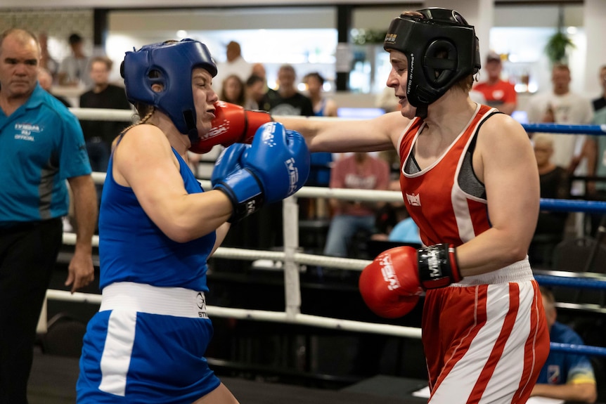Two women in red and blue sports uniforms in the boxing ring with red and blue boxing gloves and a crowd and referee behind them