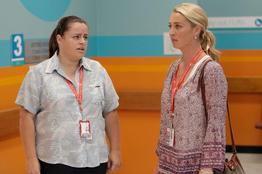 A woman with long brown hair in a pony tail stands looking at a woman with long blonde hair in a pony tail in a hospital.
