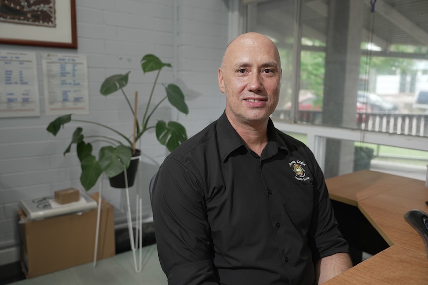 A man in a black collared shirt sits in an office.