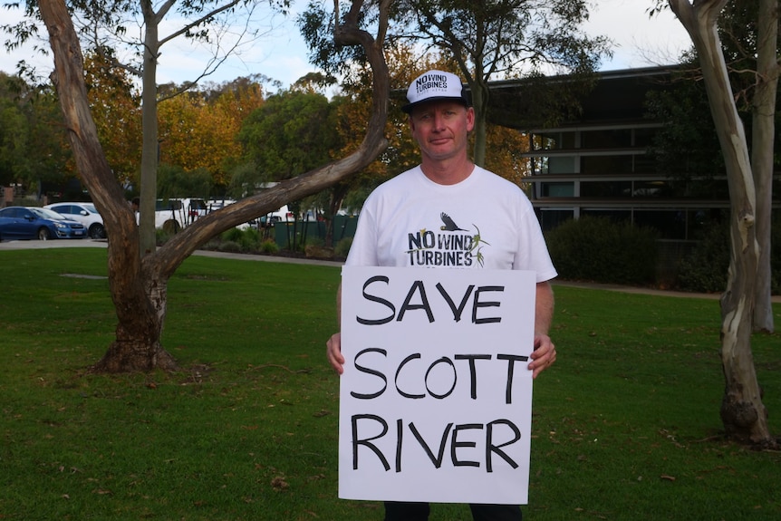 A man in a cap holds a sign saying "Save Scott River" while standing on a lawn outside a municipal building.