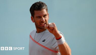 Cameron Norrie is wearing a white t-shirt with a red sash and holding up his fist