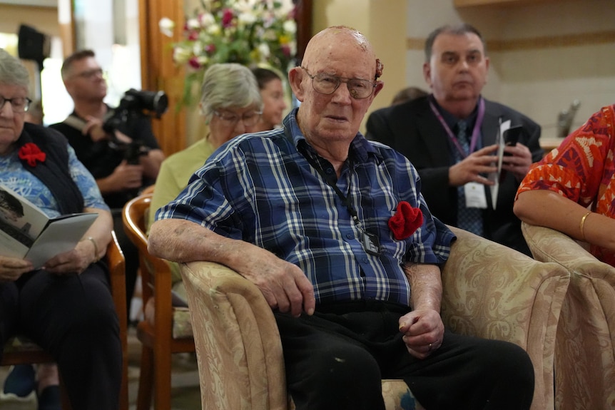 A group of elderly people sitting for a service, with one man, blue plaid shirt, looking directly at camera, sombre expression.