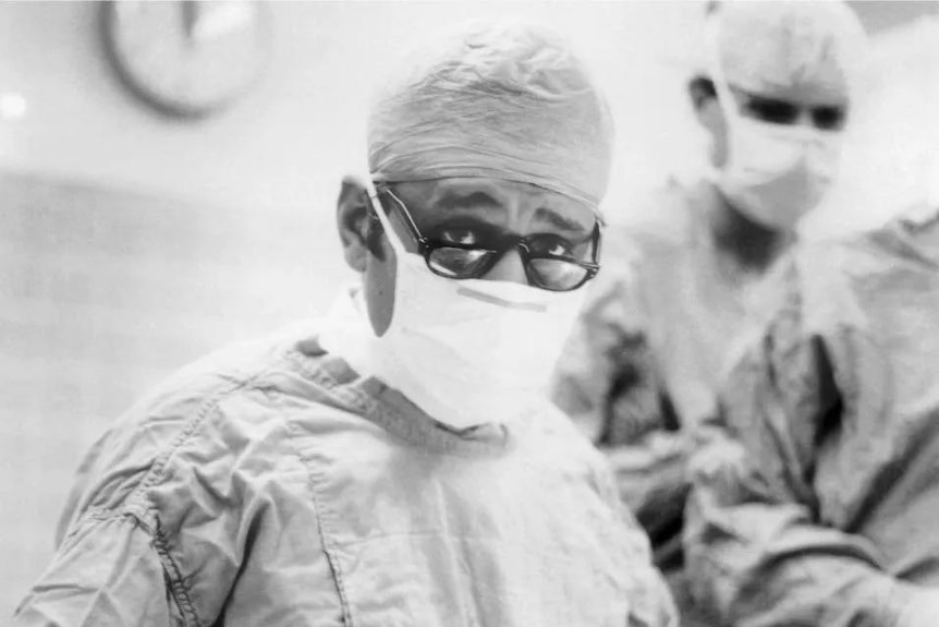 A black and white photo of a doctor in scrubs looking at the camera.