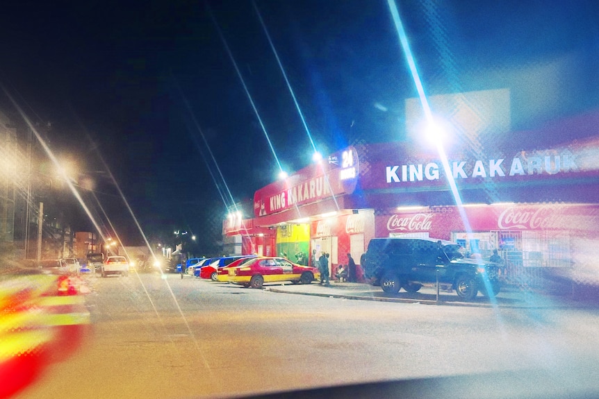 Cars sit parked outside a red shop at night with people milling on the footpath nearby