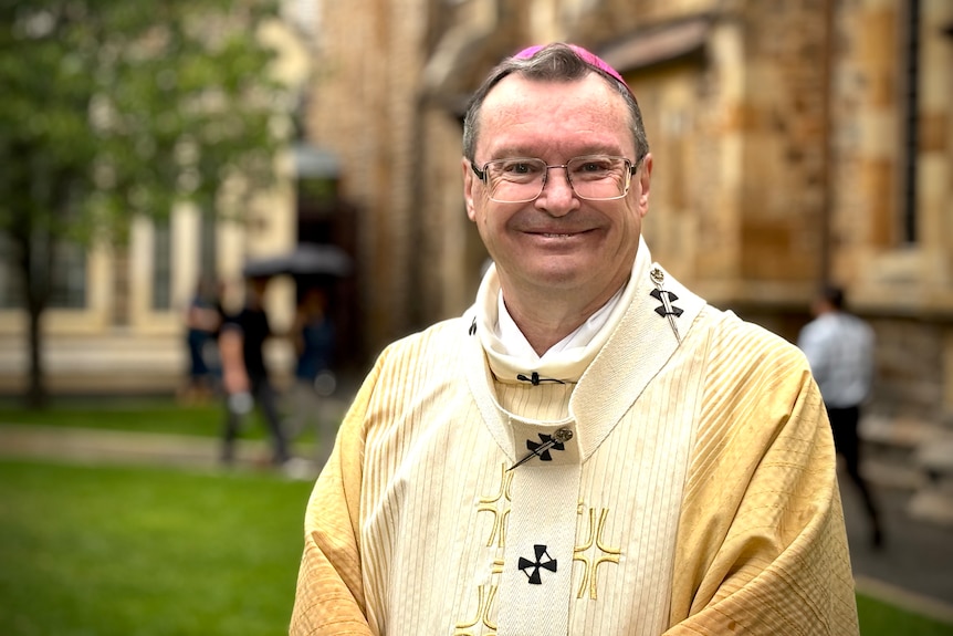 A man in gold catholic archbishop robes smiles in front of a park next to a cathedral