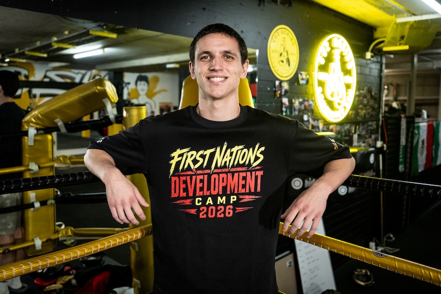 A young man with short dark hair leans against the ropes of a boxing ring smiling.