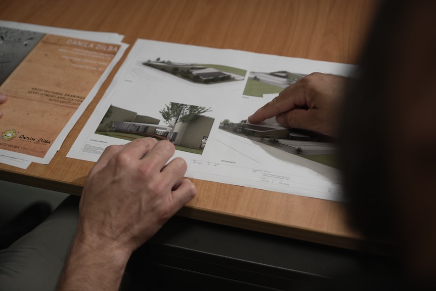 A person points at concept designs laid out on a piece of paper on a desk.