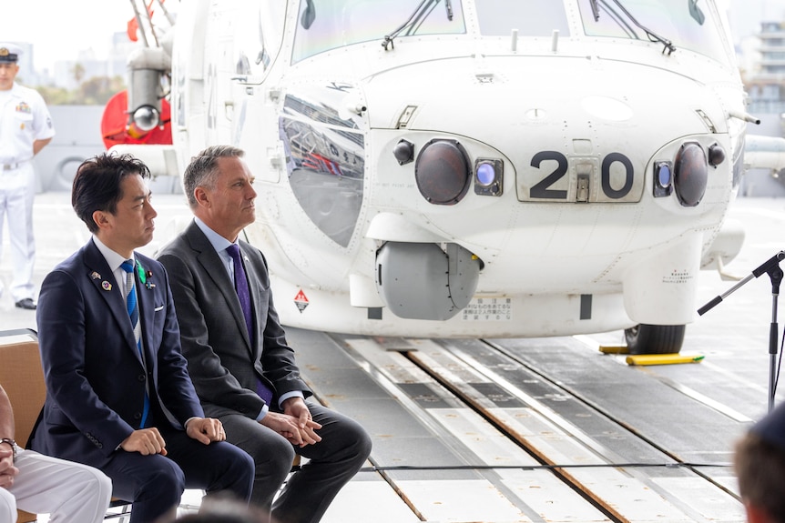 Koizumi Shinjiro and Richard Marles sit side by side in front of a warship. 