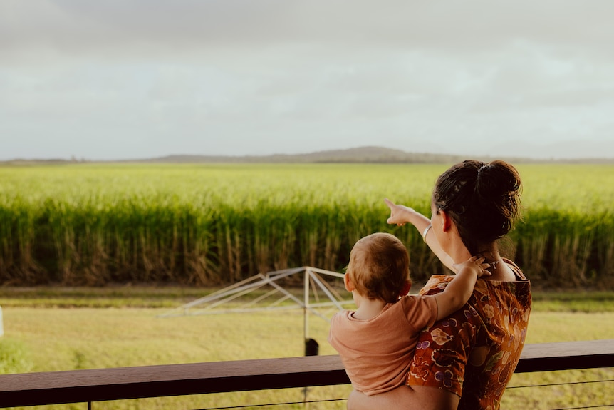 A young woman with a baby on her hip points from her verandah to a mountain overlooking a field of cane.