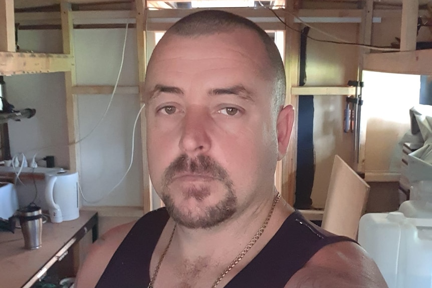 A middle-aged man with a shaved head and facial hair. He is also wearing a blue singlet and a silver necklace.
