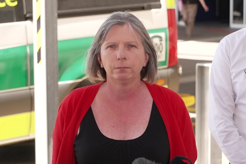 A grey-haired woman in a bright cardigan stands in front of an ambulance and speaks to the media.