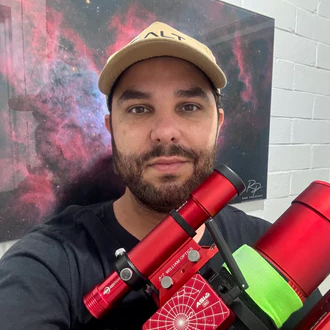A bearded man is pictured wearing a cap and dark T-shirt holding a red telescope while smiling at the camera in front of a nebula image hanging on a wall.