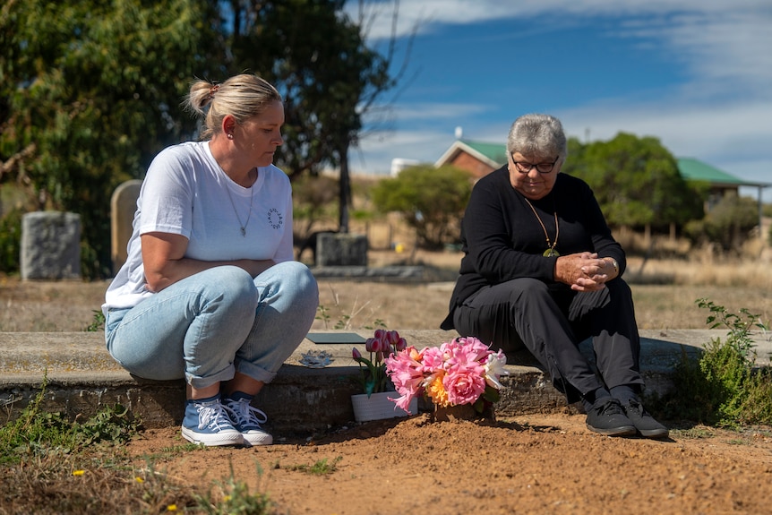 A woman in a white t-shirt and blue jeans sits alongside a woman wearing black by a grave adorned with pink flowers.