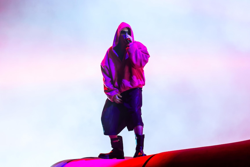 Justin Bieber in bright hooded jacket stands on a raised curved surface holding a microphone against a glowing sky