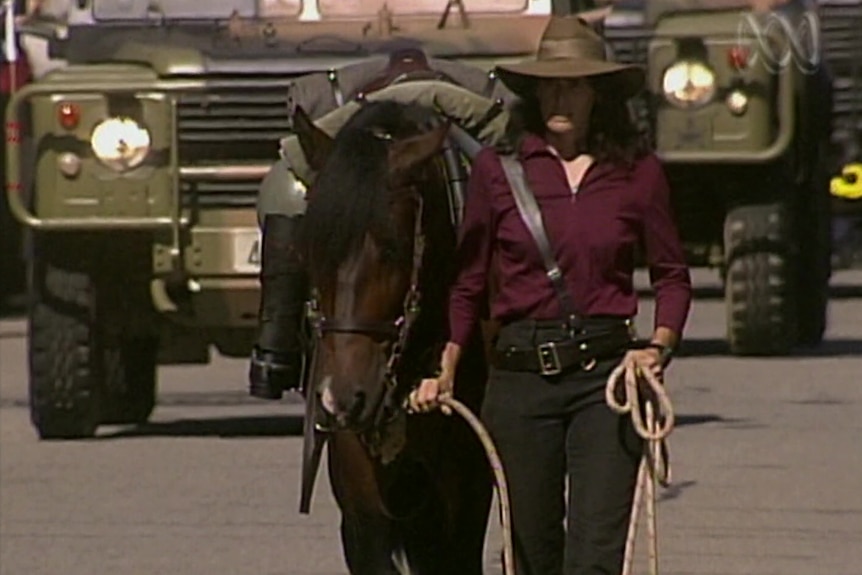 A woman wearing a wide-brimmed hat, marching next to a horse along a road in Darwin.