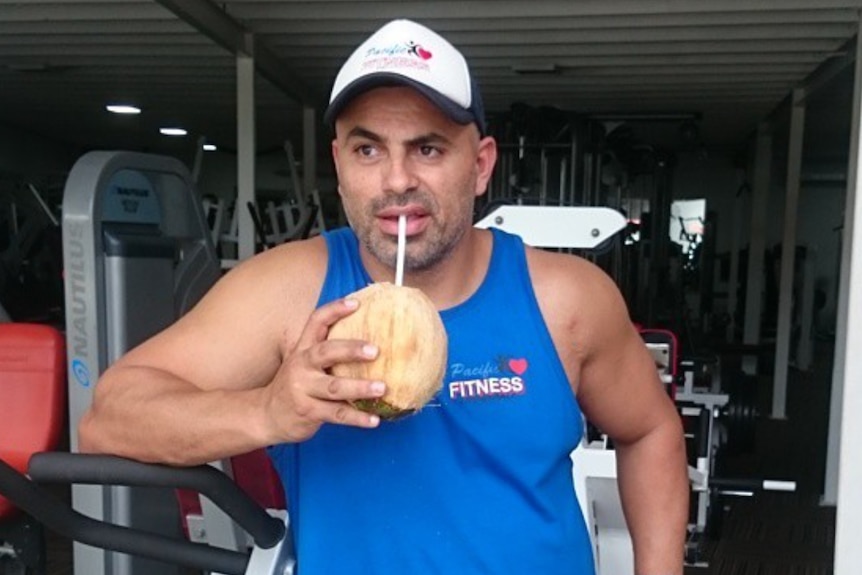 A man in a blue singlet and a baseball cap drinking a coconut through a straw with gym equipment behind him.