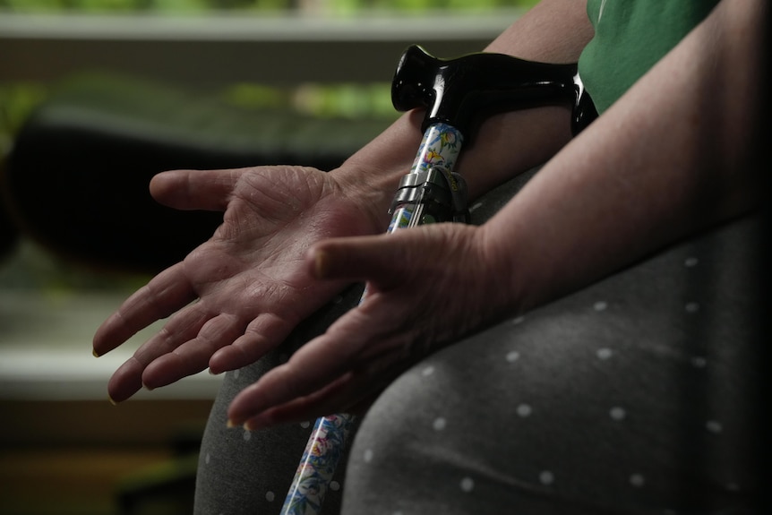 A seated woman gestures with her hands while a walking stick leans against her leg.