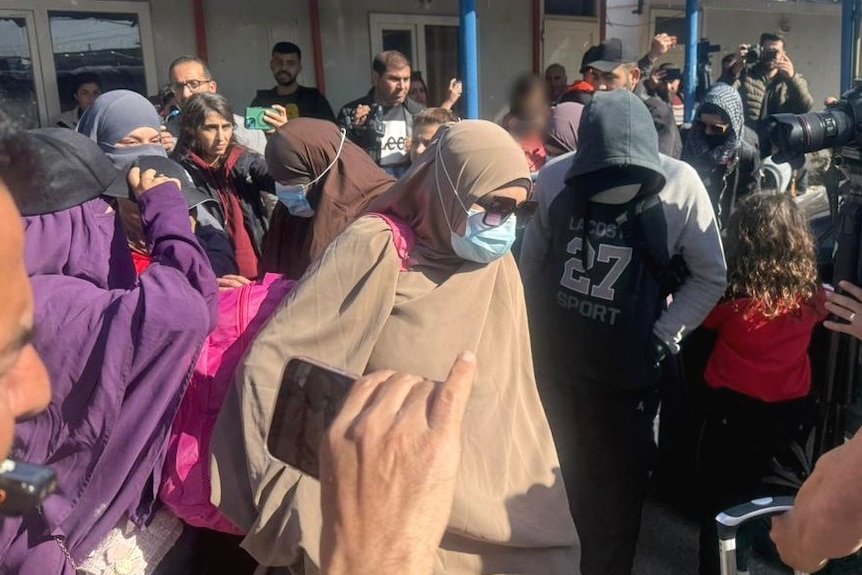 A crowd of journalists and photographers surround a small group of women wearing hijabs and face masks.