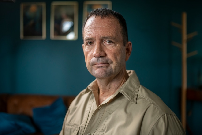 A man wearing a brown collared shirt in a mid-shot portrait standing in a room with blue painted walls