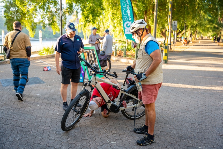A man holds his bicycle while another person inspects underneath it 
