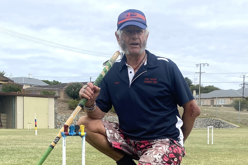 Man in cap kneeling down by hoop, with mallet and yellow, black, blue and red ball scattered in front