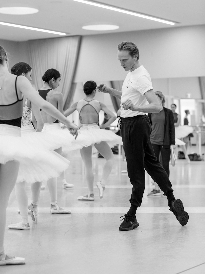 Black and white picture of David Hallberg pointing one foot in a ballet studio next to women in tutus