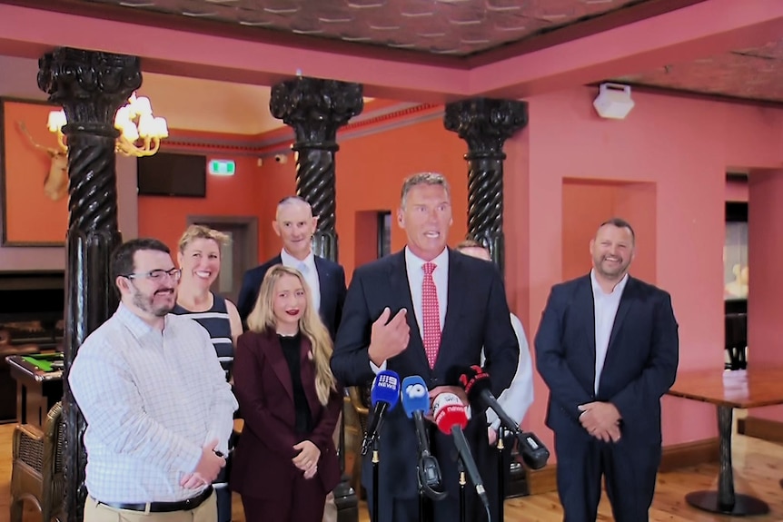 Ciry Bernardi speaks behind a group of media microphones and flanked by a group of elected One Nation members
