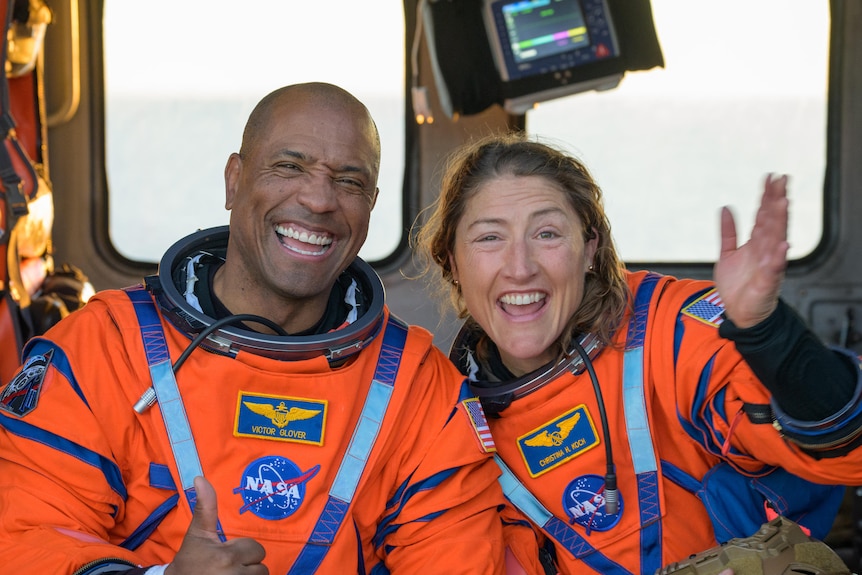 A man and a woman in orange NASA-branded space suits pose for the camera with a wave and a thumbs up