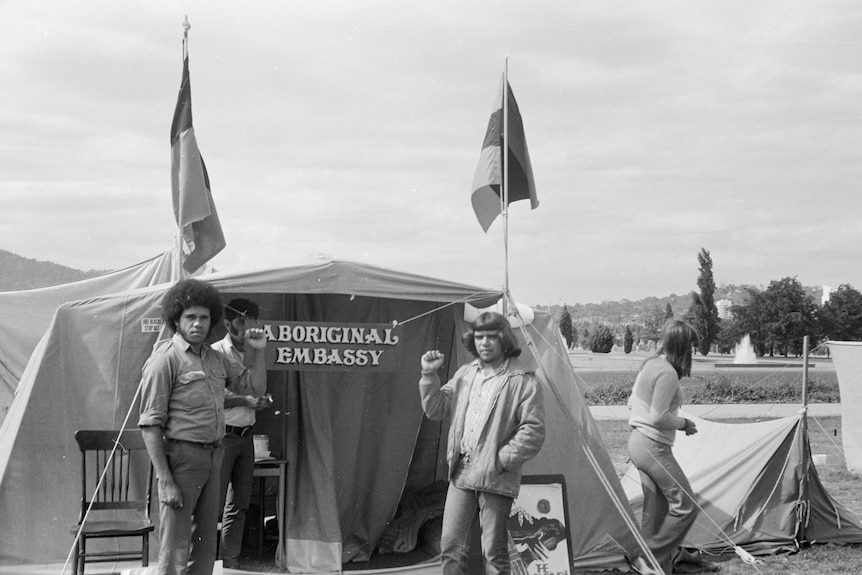 Two men stand in front of a tent that has a sign reading 'aboriginal embassy'. 